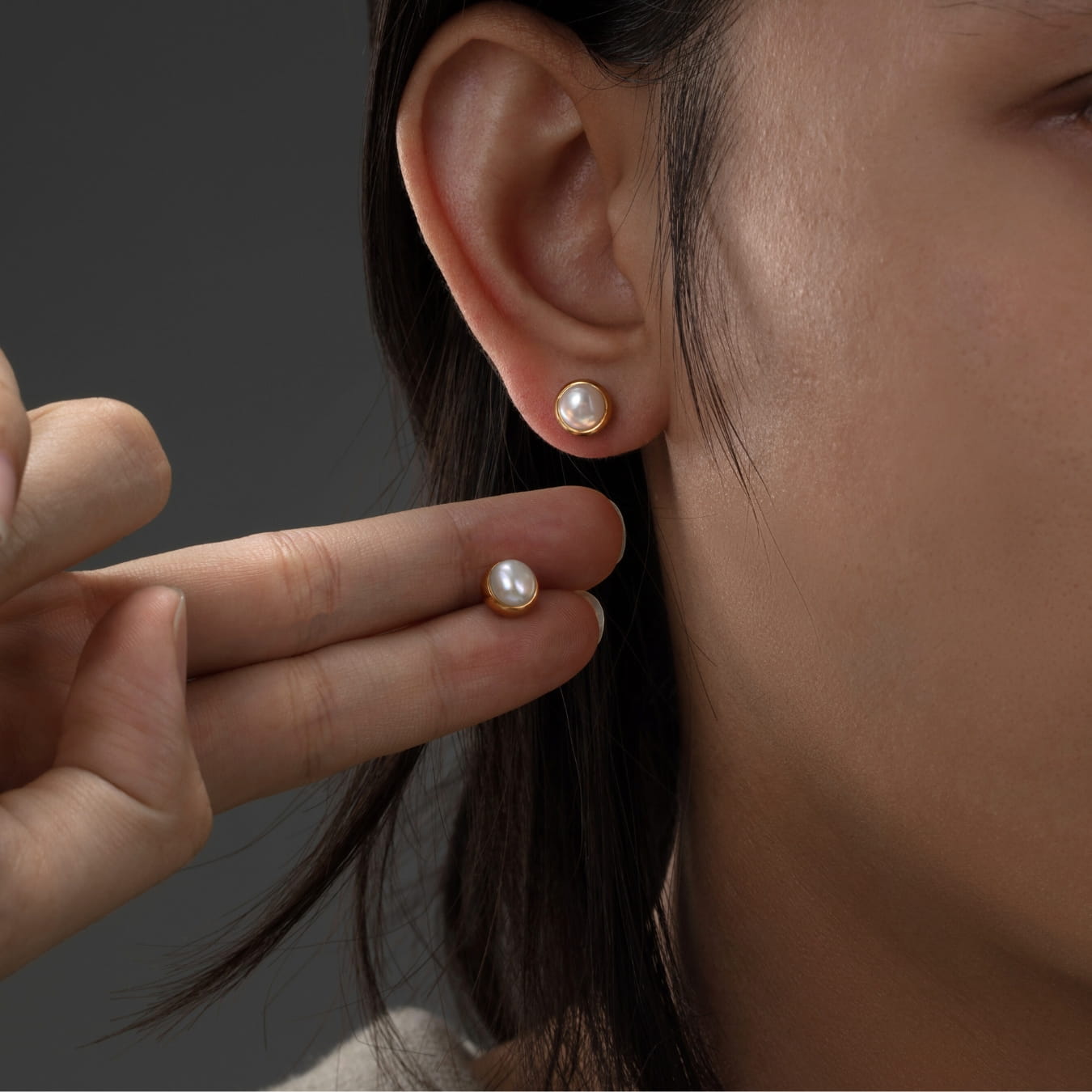 Pearl earrings being worn and held up to the ear against a dark background