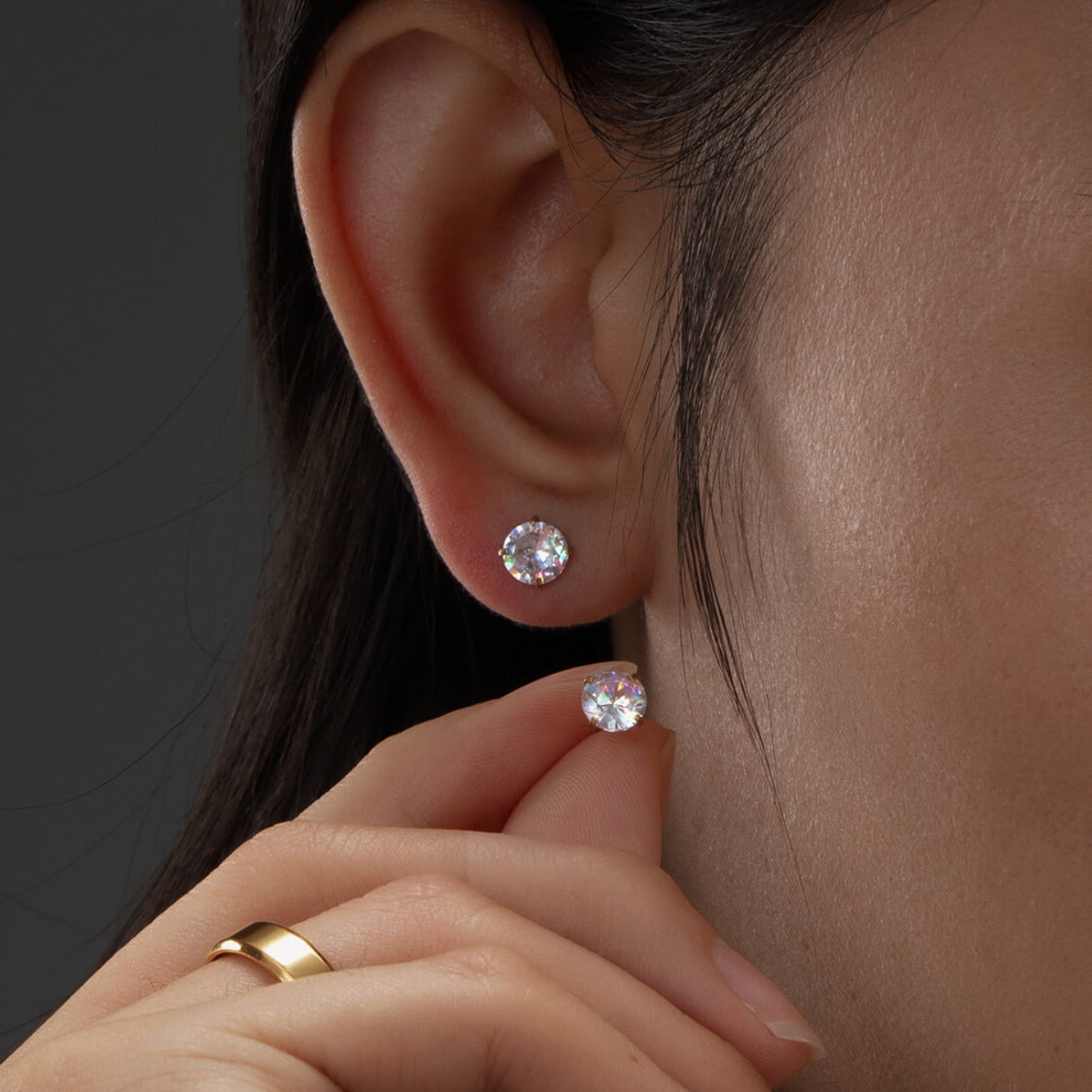 Close-up of an ear wearing two diamond earrings with a hand holding a gold ring.