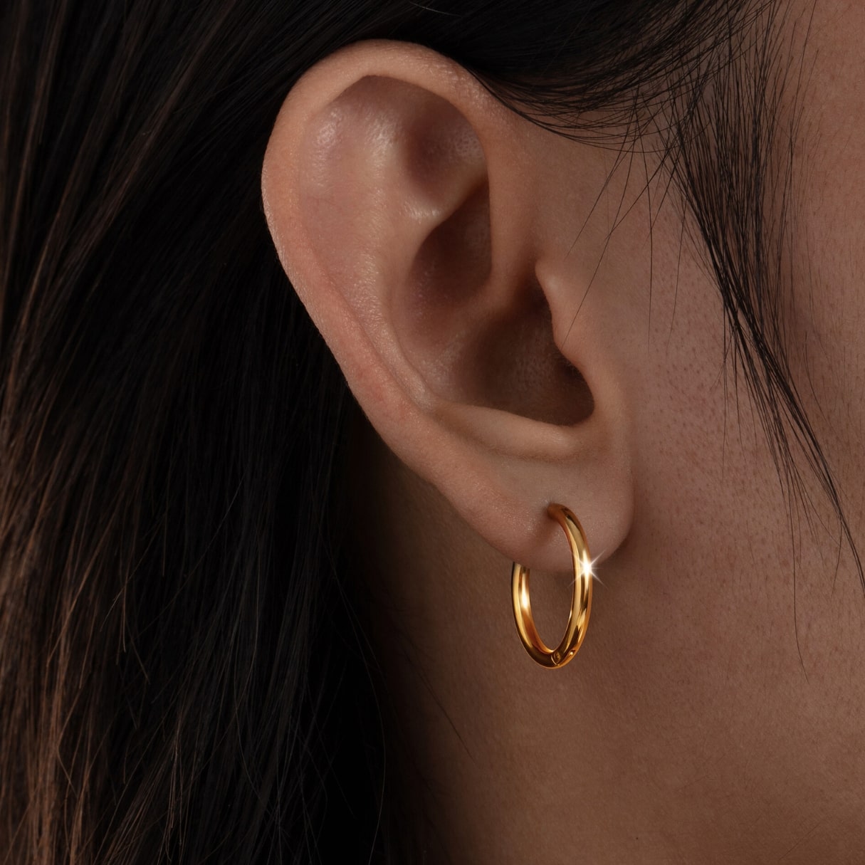 Close-up of an ear wearing a gold hoop earring with dark hair in the background