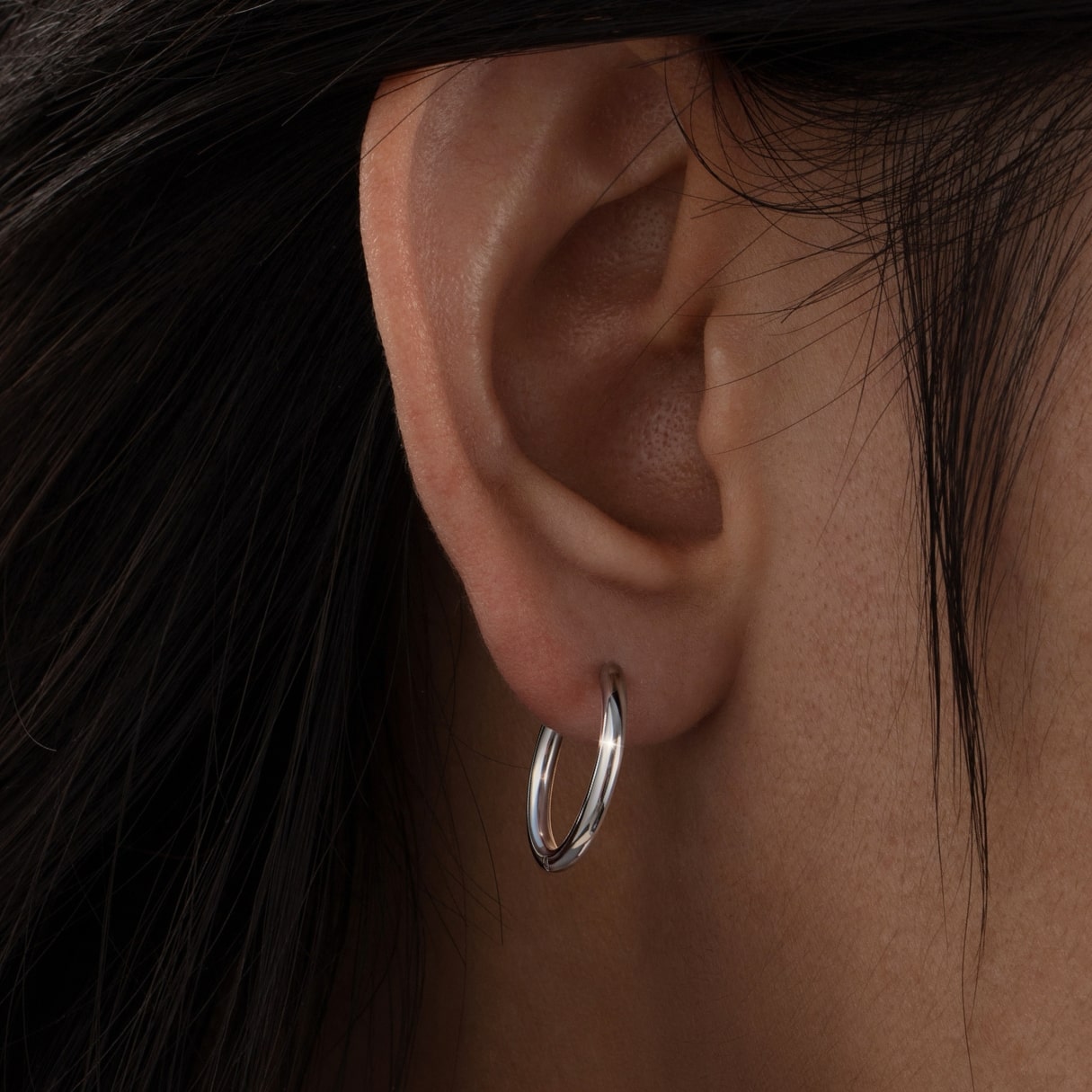 Close-up of an ear wearing a silver hoop earring with dark hair in the background